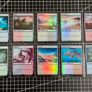May include: A collection of ten holographic Magic: The Gathering dual land cards arranged in two rows. The cards feature distinct landscape illustrations including Tropical Island, Taiga, Scrubland, Savannah, Plateau, Bayou, Badlands, Tundra, Volcanic Island, and Underground Sea, each with specific mana ability text.