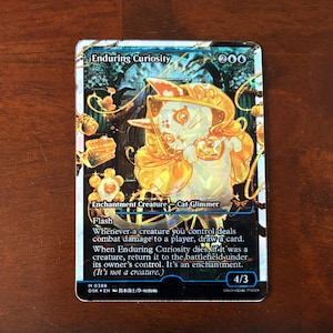 May include: A collectible card featuring a whimsical cat-like creature in golden armor, titled "Enduring Curiosity." The card has a blue and gold color scheme with text describing its abilities. The card is 4/3.