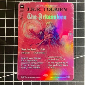 May include: A holographic trading card titled J.R.R. Tolkien The Arkenstone, featuring a bearded figure holding a glowing orb. The card text includes Seek the Heart, Sorcery-Adventure, and game mechanics. The background is a vibrant pink and orange gradient.