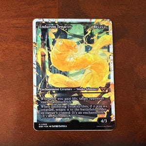 May include: A Magic: The Gathering card with a white background and black border. The card is titled "Enduring Tenacity" and features a yellow snake-like creature with a butterfly symbol. The card text reads "Whenever you gain life, target opponent loses that much life. When Enduring Tenacity dies, if it was a creature, return it to the battlefield under its owner's control. It's an enchantment. (It's not a creature)"