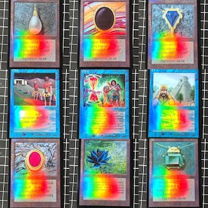 May include: Nine collectible trading cards with holographic foil effects. The cards feature various fantasy art illustrations, including a teardrop, a black gem necklace, and a blue gem pendant. Each card has text at the bottom.