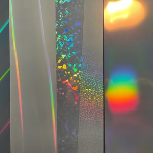 May include: Five vertical strips of adhesive vinyl film displayed side by side. The strips feature various finishes including smooth holographic gradients, a shattered glass pattern, a fine glitter texture, and a reflective surface with rainbow light.