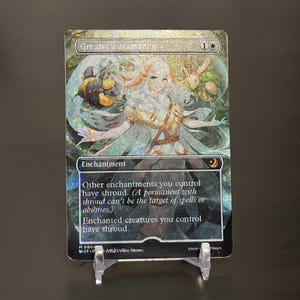 May include: A fantasy-themed trading card with a holographic finish. The card features a woman with long white hair and a white dress, holding a staff. The card text reads "Greater Auramancy 1* Enchantment Other enchantments you control have shroud. (A permanent with shroud can't be the target of spells or abilities.) Enchanted creatures you control have shroud."