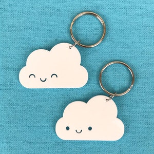 以下が含まれることがあります： 笑顔の顔をした白い雲の形をしたキーホルダーが2つ。キーホルダーにはそれぞれ銀色の金属製のリングが付いています。