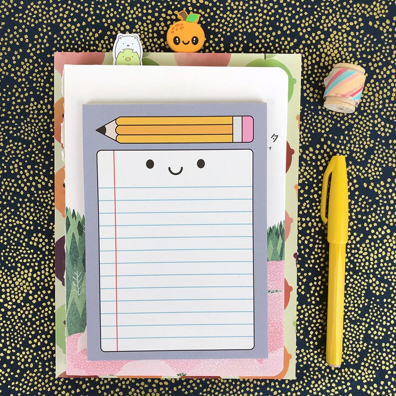 May include: A gray notepad with a smiling pencil illustration and lined paper. The notepad is on a pink and white patterned background with a small orange fruit sticker and a white bear sticker.