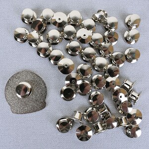 May include: A large quantity of silver-toned metal pin backs, also known as clutch backs, for use with brooches, pins, and other jewelry. The pin backs are scattered on a light gray surface.
