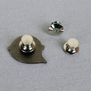 May include: The back of a silver metal pin with a black heart shape. The pin has a butterfly clasp on the back.