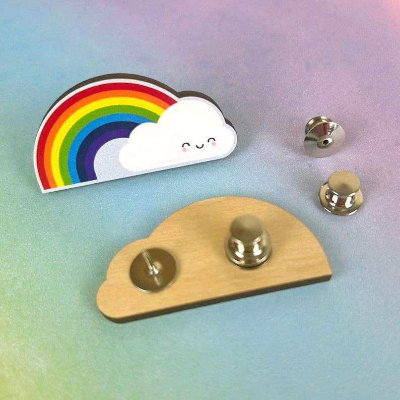May include: A wooden rainbow and cloud pin with a smiling face. The pin has a silver back with a safety pin closure.