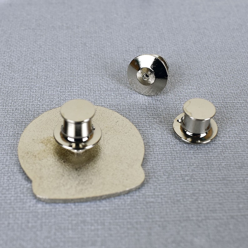 May include: Three silver metal pin backs for attaching a pin or badge to clothing. The pin backs are different sizes. The largest pin back is attached to a silver metal pin.