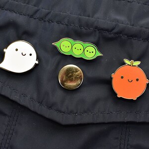 May include: Four enamel pins on a dark blue fabric background. The pins are a white ghost with a smiling face, a green pea pod with three peas, a gold circle, and an orange fruit with a smiling face.