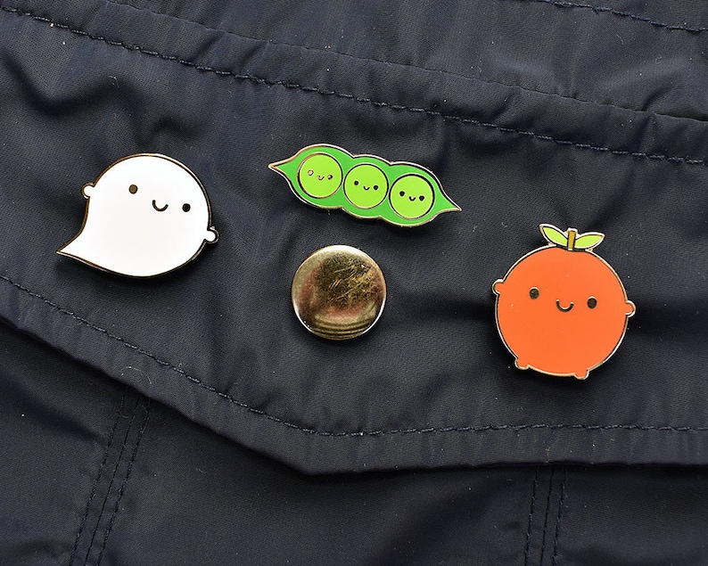 May include: Four enamel pins on a dark blue fabric background. The pins are a white ghost with a smiling face, a green pea pod with three peas, a gold circle, and an orange fruit with a smiling face.