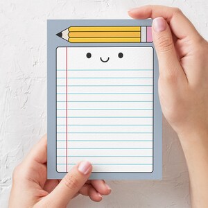 May include: A gray notepad with a yellow pencil and a happy face on the front. The notepad has lined paper and a red line down the side.