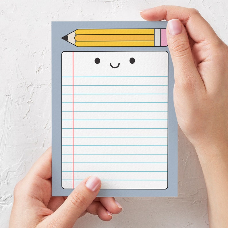 May include: A gray notepad with a yellow pencil and a happy face on the front. The notepad has lined paper and a red line down the side.