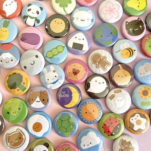 May include: A selection of colourful, round pin badges featuring charming illustrations. Designs include a panda, cat, planet, flower, and food items like an aubergine and onigiri. Some badges have text, such as "Hi There".