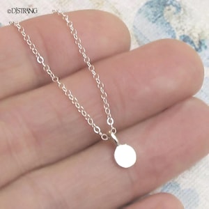 May include: A delicate silver necklace featuring a small, round pendant. The chain is fine and the pendant is a simple, polished disc. The necklace is shown against a soft background.
