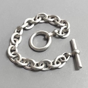 May include: A silver-colored bracelet with a chain link design. The bracelet features a toggle clasp closure. The links are oval-shaped and connected to each other. The bracelet is displayed on a gray surface.