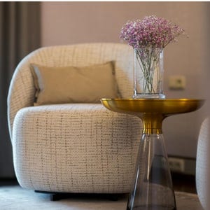 Könnte beinhalten: Ein modernes Wohnzimmer mit einem cremefarbenen Sessel und einem passenden Lendenkissen. Ein kleiner goldener Beistelltisch trägt eine Glasvase mit rosa Blumen. Darüber Stoffmuster mit der Aufschrift "Charme" in verschiedenen neutralen Tönen.