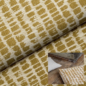 K&ouml;nnte beinhalten: Nahaufnahme eines strukturierten Stoffes mit einem sich wiederholenden Muster aus senffarbenen rechteckigen Formen auf cremefarbenem Hintergrund. Der Stoff ist gefaltet und zeigt seine Textur und sein Design. Ein Kissen mit &auml;hnlichem Muster ist sichtbar.