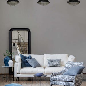 K&ouml;nnte beinhalten: Ein modernes Wohnzimmer-Setup mit einem wei&szlig;en Sofa, blauen Kissen und einem gemusterten Sessel. Ein dunkelblauer Teppich und ein schwarzer Spiegel erg&auml;nzen die Einrichtung. Der Raum wird von Pendelleuchten beleuchtet, was eine stilvolle Atmosph&auml;re schafft.