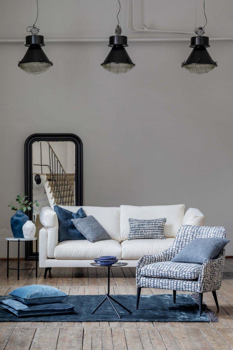 K&ouml;nnte beinhalten: Ein modernes Wohnzimmer-Setup mit einem wei&szlig;en Sofa, blauen Kissen und einem gemusterten Sessel. Ein dunkelblauer Teppich und ein schwarzer Spiegel erg&auml;nzen die Einrichtung. Der Raum wird von Pendelleuchten beleuchtet, was eine stilvolle Atmosph&auml;re schafft.