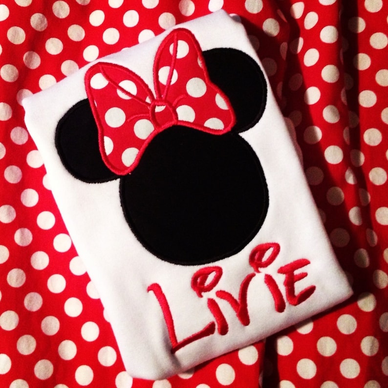 K&ouml;nnte beinhalten: Wei&szlig;es T-Shirt mit einem schwarzen und roten gestickten Minnie Mouse Kopf und dem Namen "Livie" in roter Stickerei.