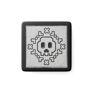 Peut inclure: Un dessous de verre carré noir avec une incrustation en tissu blanc ornée d'un motif de crâne en pixel art brodé en noir. Le crâne est centré et entouré d'un motif géométrique de petites croix et de formes en L pixelisées en fil noir.