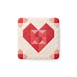 Puede incluir: Una tela cuadrada y acolchada con un diseño de corazón geométrico. El corazón está compuesto por varios tonos de rojo, sobre un fondo crema. Los bordes presentan pequeñas formas geométricas rosas. La tela tiene un aspecto cosido y texturizado.
