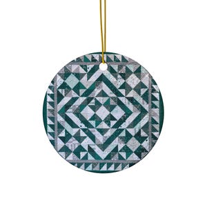 May include: A round, flat ceramic ornament featuring a geometric quilt-inspired pattern in shades of dark teal, light gray, and white. The design consists of repeating triangles and squares arranged in a central star motif with a gold hanging cord.