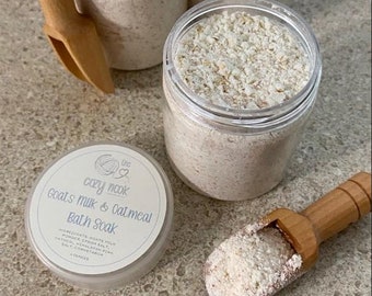 Puede incluir: Tres frascos de vidrio llenos de un baño de inmersión en polvo blanco etiquetados como "Cozy Nook Goat's Milk & Oatmeal Bath Soak". Los frascos están sobre una superficie marrón claro con dos cucharones de madera.