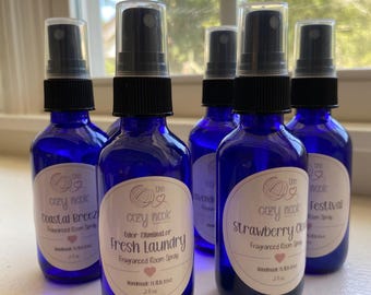 May include: Five cobalt blue glass spray bottles with black pump nozzles and white oval labels. Each label features the text The Cozy Nook, a scent name like Fresh Laundry or Coastal Breeze, and the volume 2 fl oz. The bottles are arranged in a group.