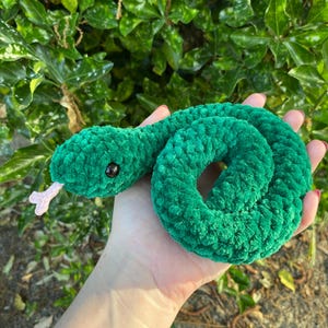 May include: A green crocheted snake toy with black eyes and a pink tongue. The snake is curled up in a circle.