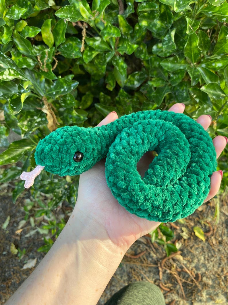 May include: A green crocheted snake toy with black eyes and a pink tongue. The snake is curled up in a circle.