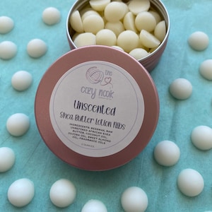 May include: A rose gold metal tin filled with small, off-white, dome-shaped shea butter lotion nibs. The lid features a label with the text: The Cozy Nook, Unscented, Shea Butter Lotion Nibs, Ingredients: Beeswax, Raw Unrefined African Shea Butter, Coconut Oil, Jojoba Oil, Sweet Almond Oil, Fragrance Oils, 2 Ounces.