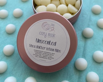 May include: A rose gold metal tin filled with small, off-white, dome-shaped shea butter lotion nibs. The lid features a label with the text: The Cozy Nook, Unscented, Shea Butter Lotion Nibs, Ingredients: Beeswax, Raw Unrefined African Shea Butter, Coconut Oil, Jojoba Oil, Sweet Almond Oil, Fragrance Oils, 2 Ounces.