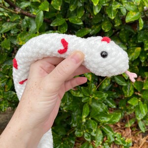 May include: White crocheted snake with red accents. The snake has a black button eye and a pink tongue.