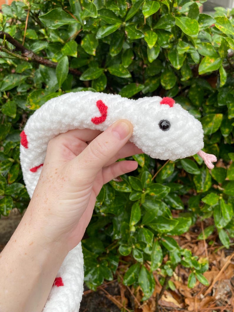 May include: White crocheted snake with red accents. The snake has a black button eye and a pink tongue.