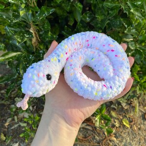 May include: A white crocheted snake with colorful confetti sprinkles. The snake is curled up in a circle and has black eyes.
