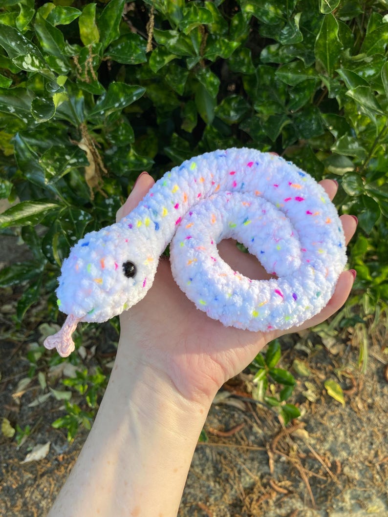 May include: A white crocheted snake with colorful confetti sprinkles. The snake is curled up in a circle and has black eyes.