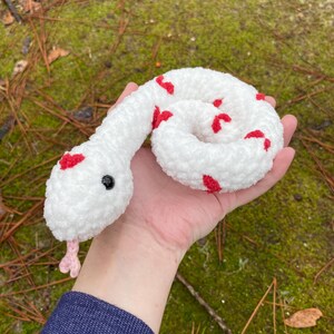 May include: White crocheted snake with red heart-shaped accents. The snake is curled up and has a pink tongue.