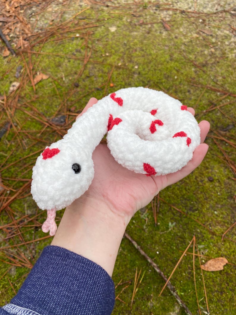 May include: White crocheted snake with red heart-shaped accents. The snake is curled up and has a pink tongue.