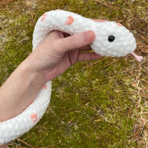 May include: White crocheted snake plush toy with pink heart accents. The snake has black eyes and a pink tongue.