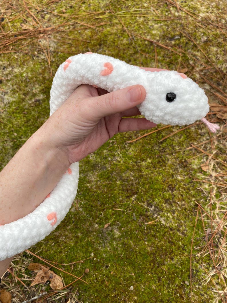 May include: White crocheted snake plush toy with pink heart accents. The snake has black eyes and a pink tongue.