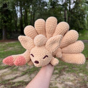 May include: A tan, plush, crocheted stuffed animal featuring a fox-like head with pointed ears, a dark brown nose, and closed eyes. The creature has nine large, rounded, fan-like tails extending from its back and two front paws with pink tips.