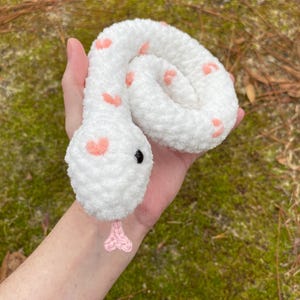 May include: White crocheted snake with pink hearts and black eyes. The snake is curled up in a hand.