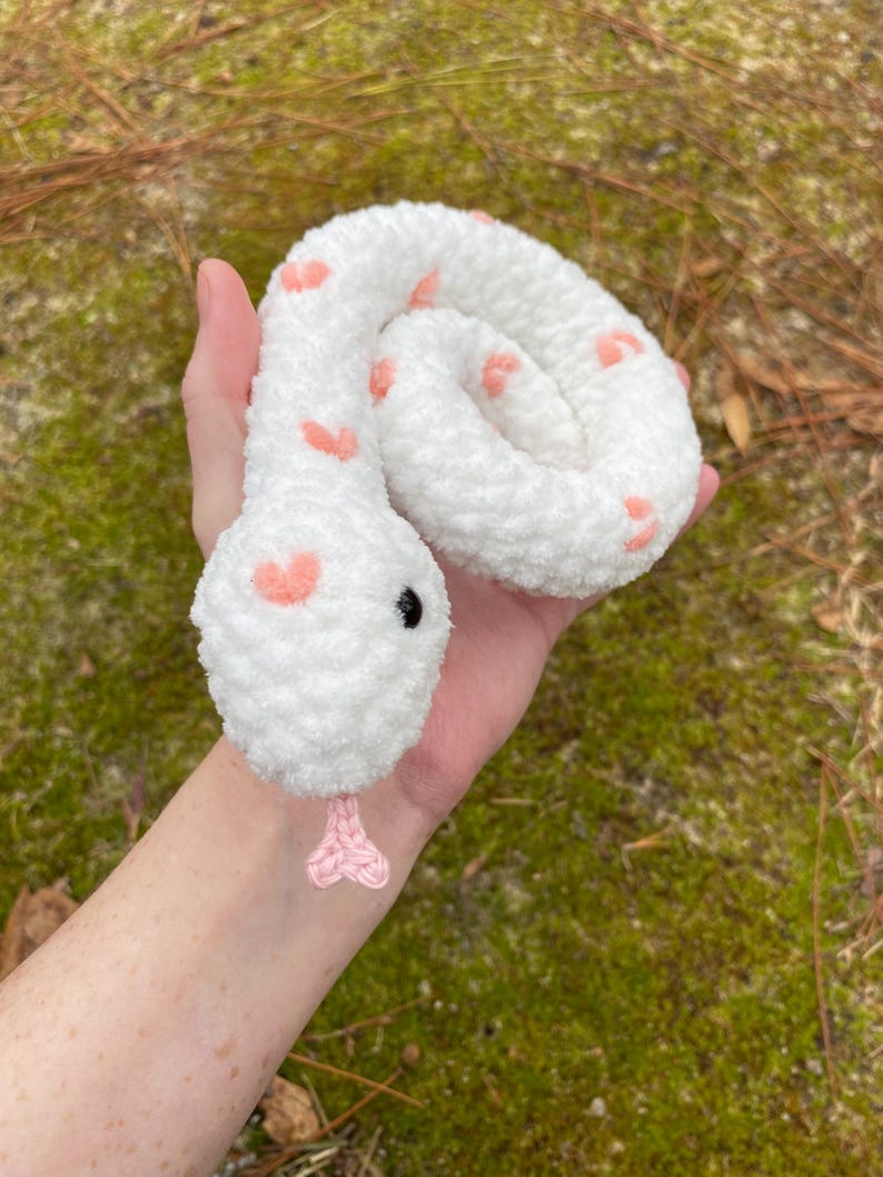 May include: White crocheted snake with pink hearts and black eyes. The snake is curled up in a hand.