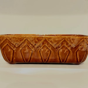May include: A rectangular brown ceramic planter with a glossy finish. The planter features a repeating geometric pattern of diamond shapes along its sides. The planter is designed for indoor or outdoor use.