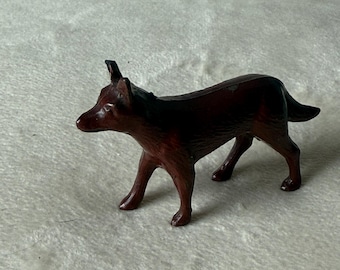 May include: A small, brown figurine of a dog in a standing pose. The dog has a pointed snout, upright ears, and a curved tail. The figurine is made of a smooth, glossy material.