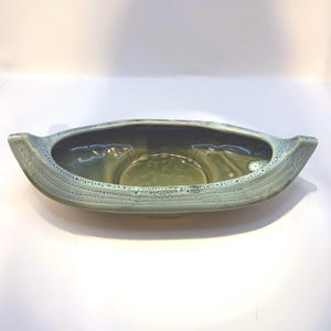 May include: A ceramic, boat-shaped planter in shades of green. The planter has a textured exterior with a speckled, light green rim and a darker green interior. It features handles on each end.