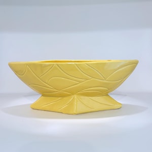 May include: A yellow ceramic planter with a leaf pattern. The planter has a rectangular shape with a wide opening and a pedestal base. The surface has a glossy finish.