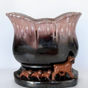 May include: A ceramic vase with a flared, scalloped rim. The vase transitions from a dark brown base to a pink and brown mottled top. Brown dog figurines encircle the base, with a larger dog leading three smaller pups.